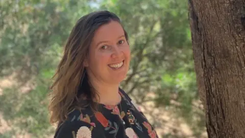 Supplied Dr Emma Lawrance standing beside a large tree wearing a dark floral-patterned top. She has long brown hair and is smiling. The scene is outdoors in a wooded area.