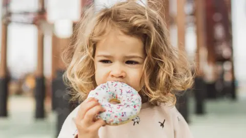 Getty Images The image shows a young child outdoors holding a large doughnut with white icing and colourful sprinkles. The child is taking a bite from the doughnut, which occupies much of the foreground. The background contains blurred structural elements, such as columns or beams, creating a shallow depth-of-field effect. Soft natural lighting highlights the child's curly hair and the pastel tones of the clothing and doughnut icing.