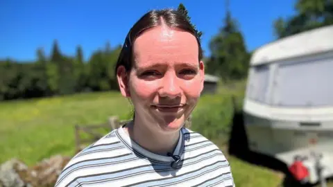 Samantha Donaldson has dark hair in a low ponytail. She is smiling at the camera into the sun with a caravan and grassy field blurred in the background.