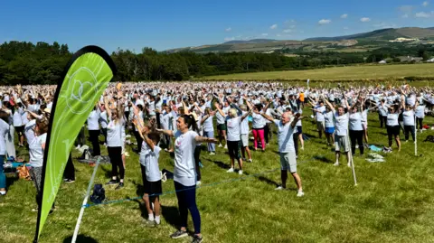 A large group of people stand gathered in a field with their arms upstretched, wearing white pilates t-shirts.
