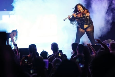 Lizzo in a skimpy black outfit playing a flute on stage with smoke behind her