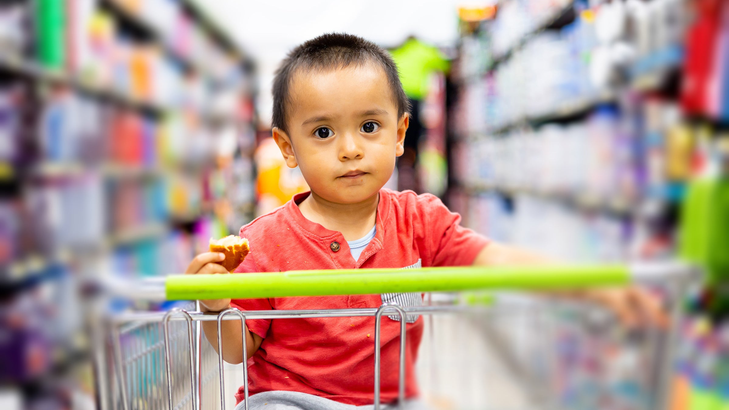 A toddler in a grocery cart holds a bread roll that has been partially eaten, shelved goods on the grocery aisle around the child.