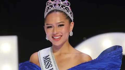 Lee Dare Angela Aumonier has dark hair tied back, is wearing a blue ball gown, a white sash over the shoulder where you can see the word 'miss' in black and is wearing a jewelled tiara.