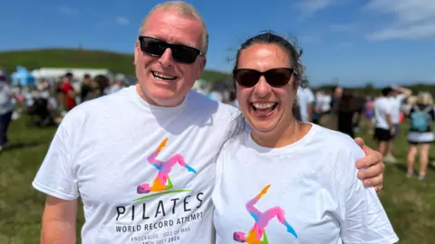 BBC Simon and Karen are standing together in the sunshine smiling wearing white Pilates t-shirts with an illustration of a Pilates move recreating the island's triskelion.