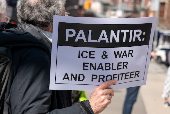 Someone holding up a sign at a protest that says "Palantir: ICE & War endabler and profiteer"