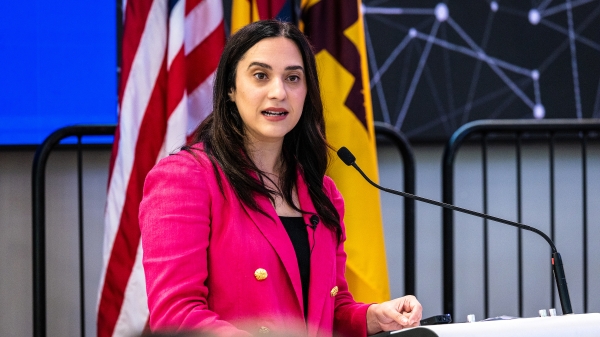 Woman with dark hair wearing a hot pink blazer speaks into a microphone to an unseen audience.