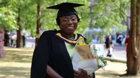 University of Manchester Makagbeh has dark hair and green eyeshadow. Smiling for the camera, she wears a green dress and a black mortar board cap and gown.