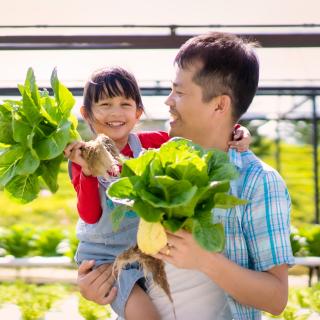 A man holds a little girl with one arm and they both hold a head of lettuce in one hand, with a farm in the background