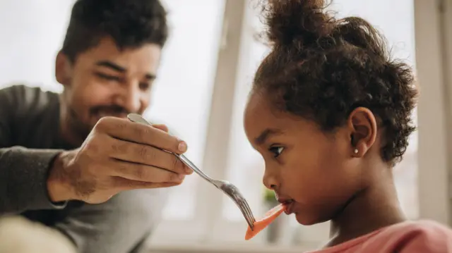 A father dey feed im daughter piece of carrot with a fork. She sidon for di foreground and dey reluctant, wearing a pink t-shirt.