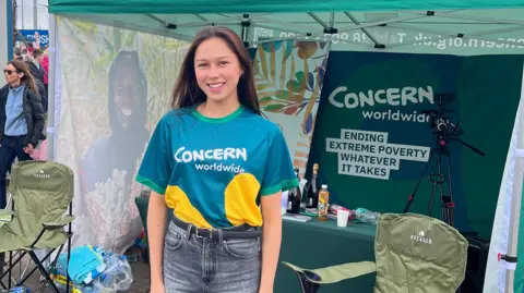 Angela Aumonier Angela Aumonier is wearing a blue "Concern Worldwide" t-shirt and standing in front of a charity tent. She has long brown hair and is smiling.