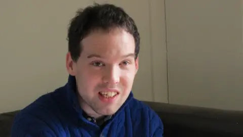 Steph Morris Tom Wentworth is pictured in a blue knitted jumper sitting indoors on a dark sofa, facing the camera. The image is a close-up portrait with a plain cream-coloured wall in the background and soft natural lighting.