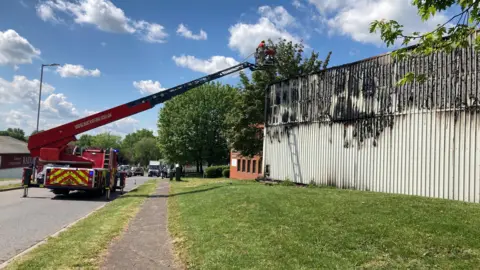 Alex Pope/BBC The outside of a building that has been burnt, with a fire engine with a crane and people on it. There is grass around the area and trees in the distance.