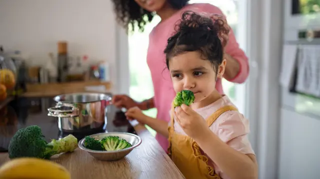 A little girl stand next to a kitchen counter as she dey smell a piece of broccoli.
