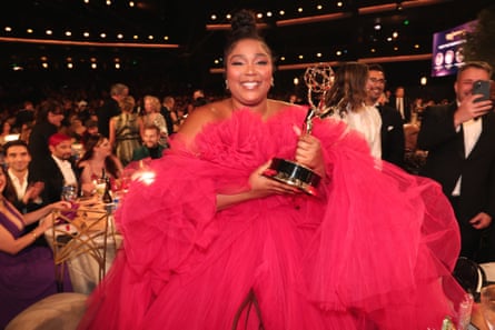 Lizzo wearing a massive pink netted outfit, holding an award and smiling at the camera. People applause in the background