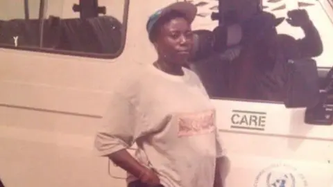Makagbeh Princess Sesay Woman wears a large t-shirt and cap standing next to a van which says 'CARE' on the side. A man is in the van.