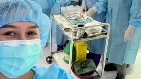 Anglea Aumonier Angela is wearing a blue face mask, hair net and blue paper overalls. Behind her is a silver trolley with medical equipment, and a group of others dressed the same.