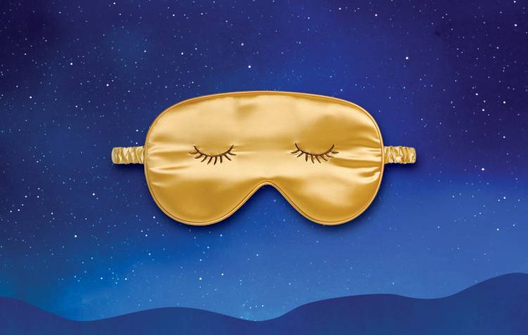 Golden satin sleep mask with embroidered closed eyes against a blue, starry night-sky background.