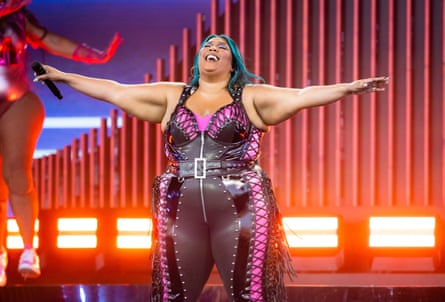 Lizzo performing on stage, standing with her arms outstretched, wearing a pink and black skintight outfit