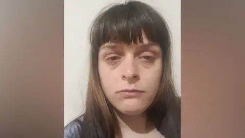 Abi Smith A selfie of a woman with long brown hair and a fringe over her forehead. She is not smiling and has dark bags under her eyes.