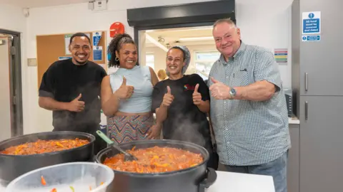 City of Wolverhampton Council Three men and a woman stand behind two big cooking pots of food on a table in a kitchen. All four of them have their thumbs up and are smiling at the camera. There is a a large kitchen hatch behind them