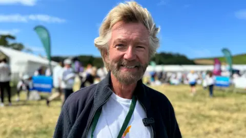 Noel Edmonds has wavy fair short hair and a short beard. He is smiling, wearing a white t shirt and woolly hacket, standing in a field with crowds behind.