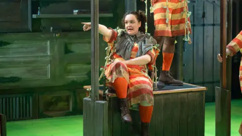 Lloyd Evans Daneka Etchells performs in a red-and-orange striped costume. She is sitting on a wooden platform and pointsingacross a theatre stage. Other cast members in matching costumes are partially visible among dark wooden set pieces under green stage lighting.
