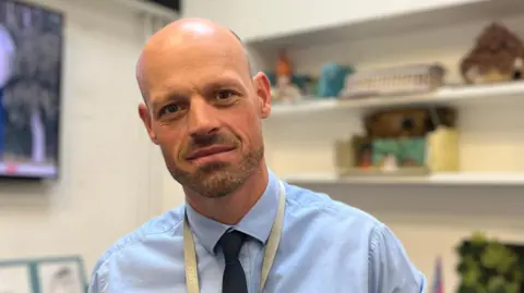 BBC A man in his 30's with a bald head and slight facial hair wearing a blue shirt with a navy blue tie