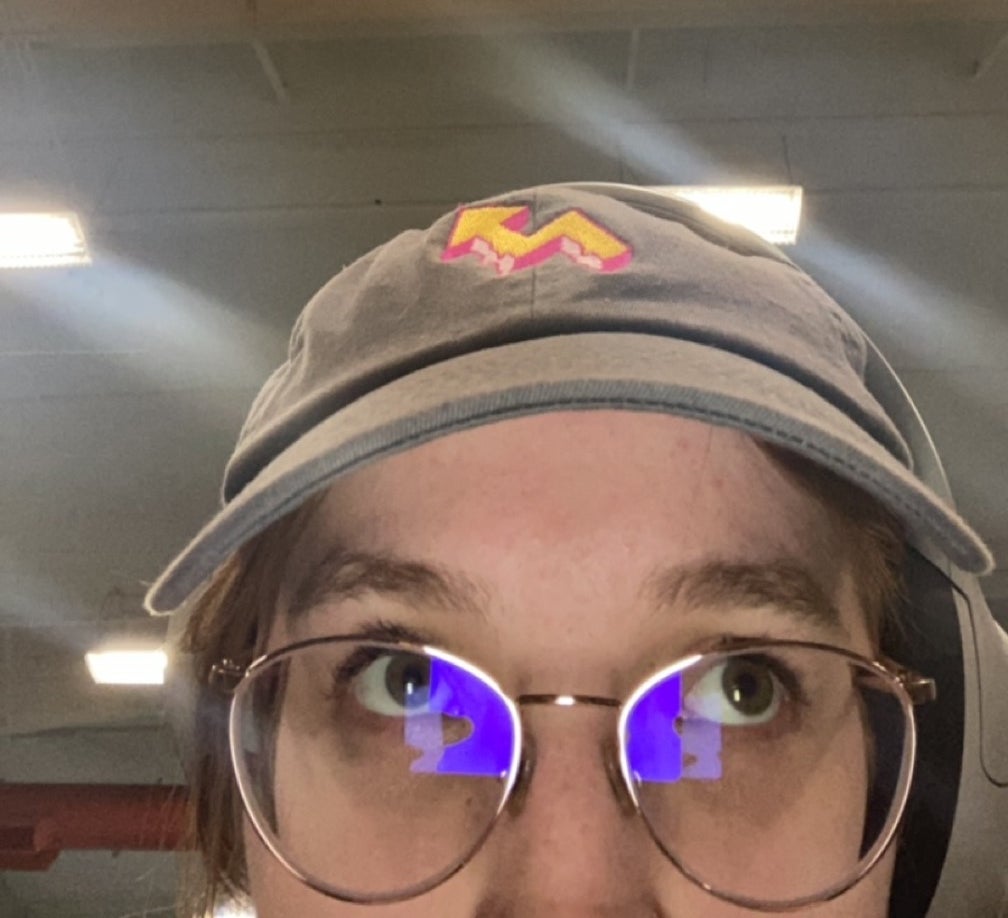 Person wearing glasses and a cap is looking upward in an indoor setting with overhead lights and ducts visible