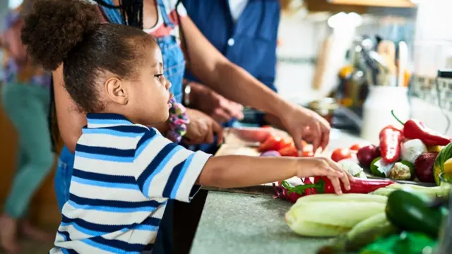 A small boy with im hair tied back dey touch a red pepper on a kitchen counter, while a woman dey cut vegetables next to am.