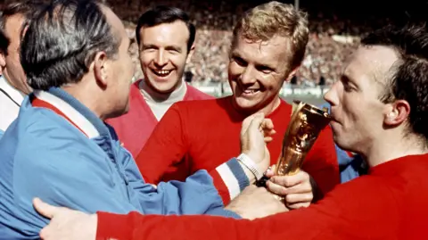 PA Media Sir Alf Ramsey in a light blue tracksuit (far left), Jimmy Armfield (centre left) with black hair in light turtle neck shirt and red sweater, Bobby Moore (centre right) with a red England shirt has blonde hair and Nobby Stiles (far right) has balck hair and  red England shirt.
