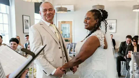 Supplied Lee, a man in a beige suit, holding Kacey's hands, who is a women in a white wedding dress. They are standing in front of friends and getting married.