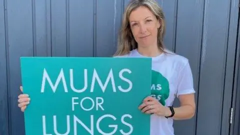 Liz Godfrey Liz Godfrey is standing in front of a blue paneled wall and she is holding a large turquoise sign reading with white lettering which reads: 'MUMS FOR LUNGS' in capital letters.