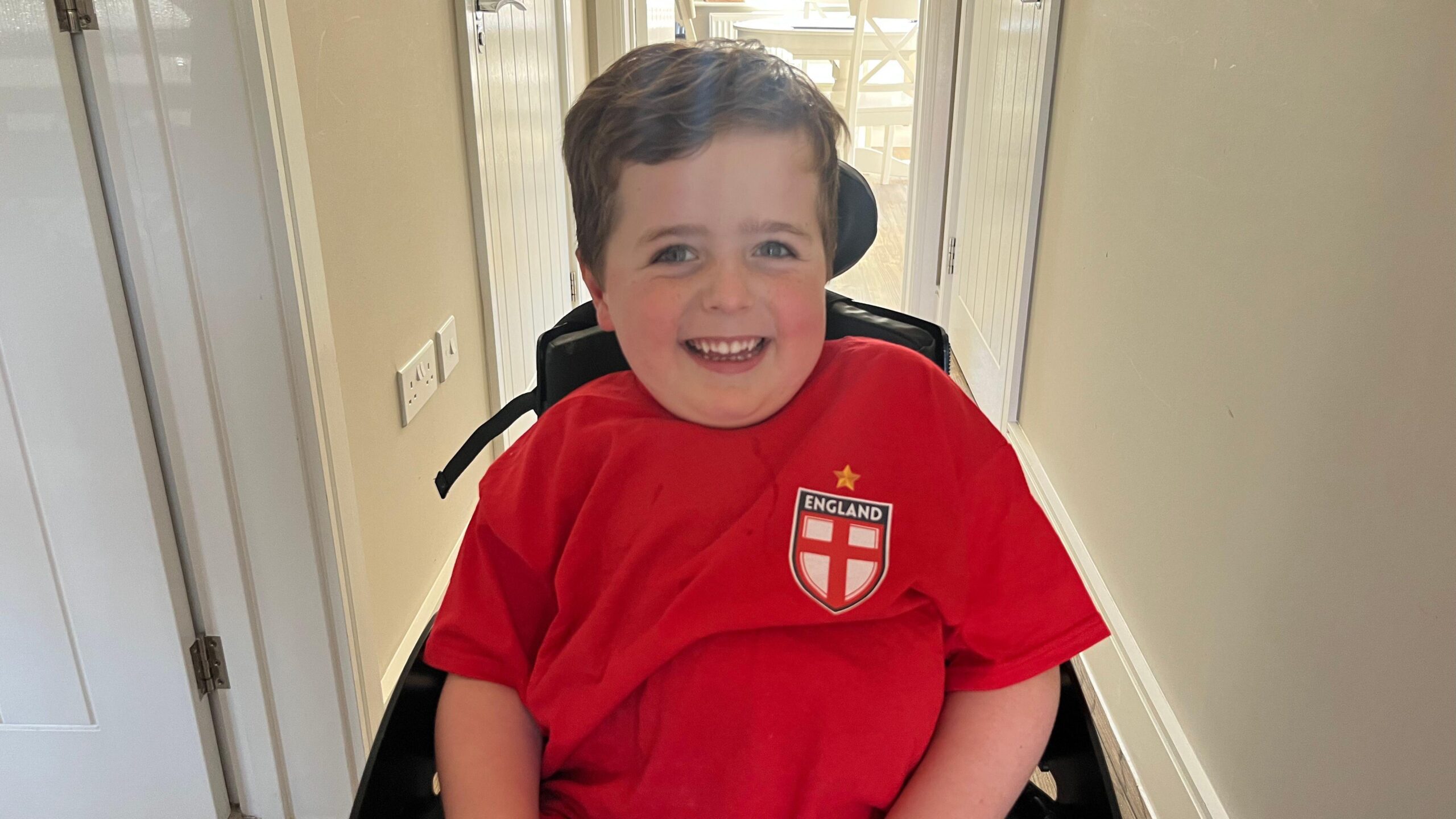 A young boy in a red England top sits in a wheelchair in a house.