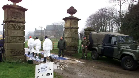 PA Media A soldier guards the entrance to a farm as three people in white hazmat suits walk onto the premesis.