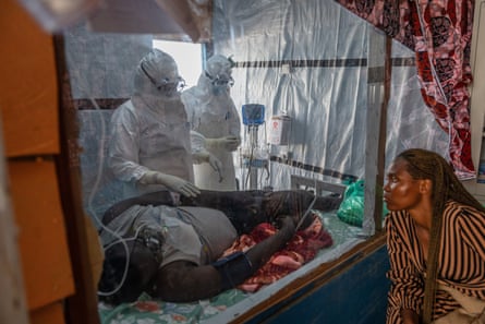 Health workers dressed in PPE tend to a man suffering from Ebola who is lying on his side, as a relative watches on from the other side of a sheet of glass