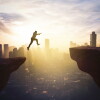 Young businessman jumping through gap cliff at sunrise time with modern city background