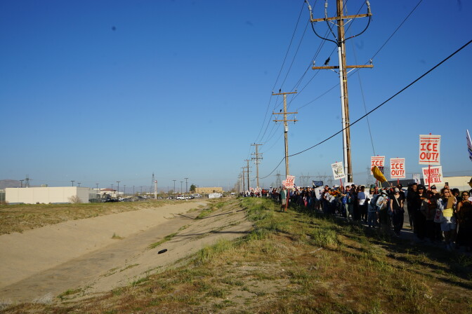 A long line of protester carrying signs that read "ICE OUT!" stand under power lines opposite a detention center on the other side of a concrete drainage channel.