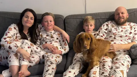 Sophie Fisher Four people are seated together on a grey sofa wearing matching festive-print pyjamas, creating a coordinated family portrait. A brown dog stands in the foreground, positioned between the seated people and looking directly at the camera.