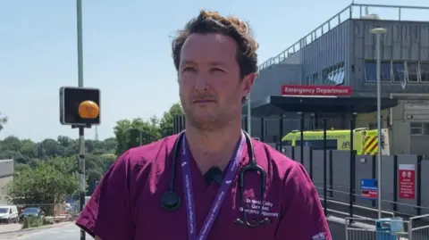 A man called Dr Ian McCarthy stands in front of Derriford Hospital in Plymouth. He has purple medical scrubs on and a black stethoscope round his neck. He is stood near a zebra crossing and an ambulance is parked behind him.
