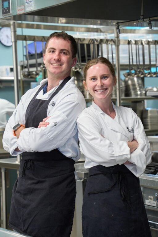 A man and a woman, both wearing chef's coats and aprons, stand together in a kitchen.