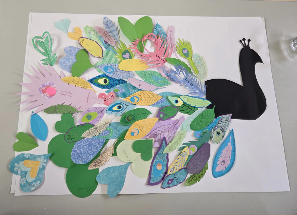 A paper peacock with a black silhouette body and colorful tail feathers made from various decorated paper cutouts, including hearts and feather shapes, displayed on a white background.
