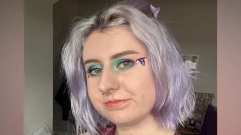 Abbie Filer A headshot of a young woman with short light purple hair. She has blue and purple eyeshadow with a uterus-shaped sticker in the corner of her eye, and and a silver septum nose piercing.