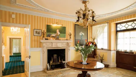 Kings West Estate Agents The entrance hall of Moulton Grange features an elaborate gold and black Baroque-style chandelier, hanging above a polished wooden table with a vase containing red and white lilies. The wallpaper is striped yellow above a white dado rail, and below it is yellow painted panels. The white stone fireplace has highly ornamental decoration surrounding a black grate. Above the fireplace hangs a painting of an older man relaxing in a chair in the garden.