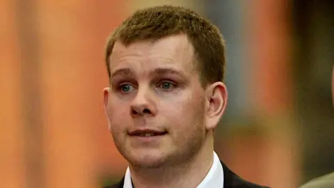 Spindrift Shaun McKenna has short, brown hair. He is wearing a white shirt and a dark suit jacket. He is staring straight ahead