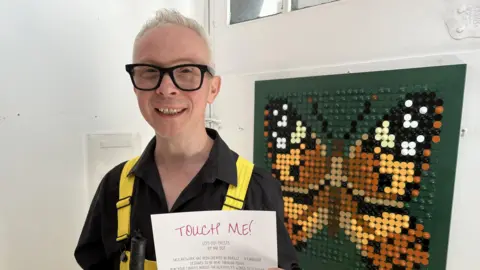 Leanne Rinne / BBC A man, Clarke Reynolds, holding a piece of paper reading "Touch Me!" in front of a butterfly art design made up of lots of dots.