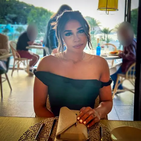 Ravika A photo of a young woman sitting at a table in a busy restaurant. Her brunette hair is tied back and she is wearing an off-the-shoulder black top, with her nude-toned handbag on the table.