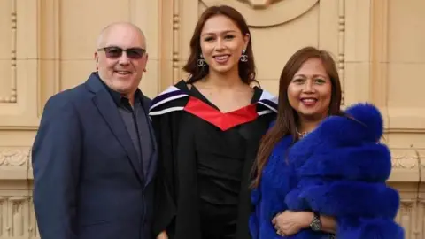 Angela Aumonier Angela is standing in black and red graduation robes. To her left is her step dad David Harris, who is wearing a navy suit. And her mum Angie Harris on the right with long dark hair and a fluffy blue jacket.