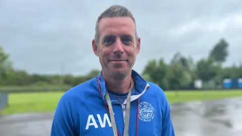 A man with short grey hair wearing a blue sports long sleeved top with the logo AW in white writing with a red whistle around his neck