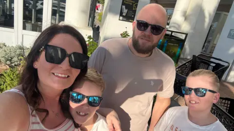 Sophie Fisher A sunny outdoor selfie shows four people wearing sunglasses gathered close together outside a modern building with white columns and large windows.
Two adults and two children are visible in the foreground, dressed casually in light-coloured summer clothing, with landscaped plants and paved walkways surrounding them.