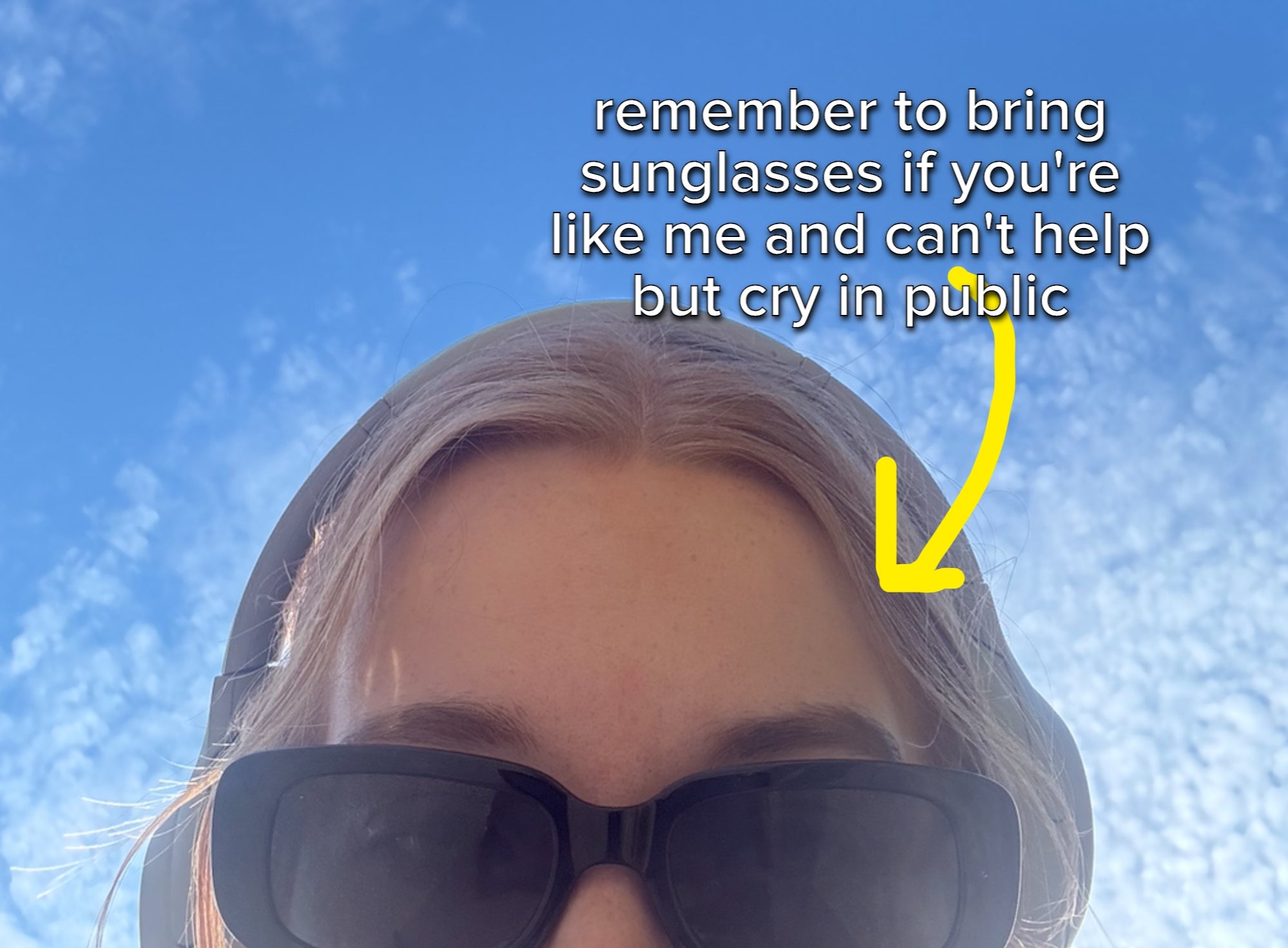 Person with headphones and sunglasses looks up towards a partly cloudy sky with tree branches above